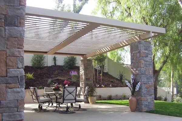 Backyard view of a house with a white pergola over a patio set, next to a green lawn under a blue sky.