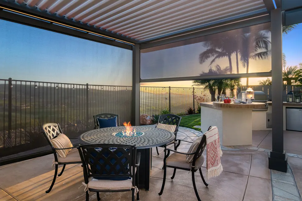 Outdoor dining space with round fire pit table under a pergola with retractable screens at sunset.